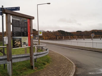 Eine Informationstafel an einer Straße vor einer Brücke über einen See im herbstlichen Landschaftsumfeld.