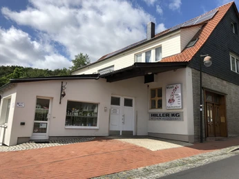 Eine helle Fassade mit einem Schild "Hiller KG", vor einem Geschäft mit gepflastertem Zugang.