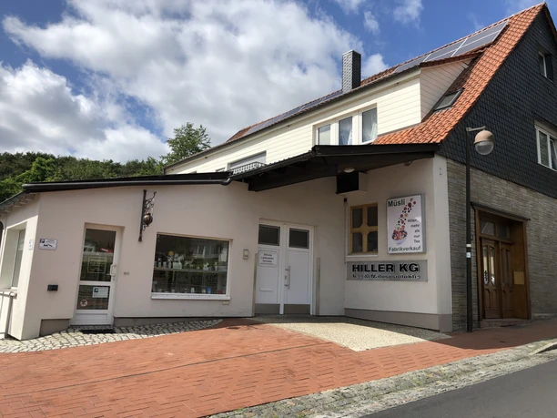 Eine helle Fassade mit einem Schild "Hiller KG", vor einem Geschäft mit gepflastertem Zugang.