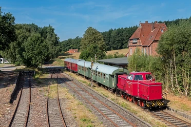 Heide-Express Rote Diesellok zieht grüne und rote Waggons auf Schienen vorbei an Backsteinhaus und grünen Bäumen.
