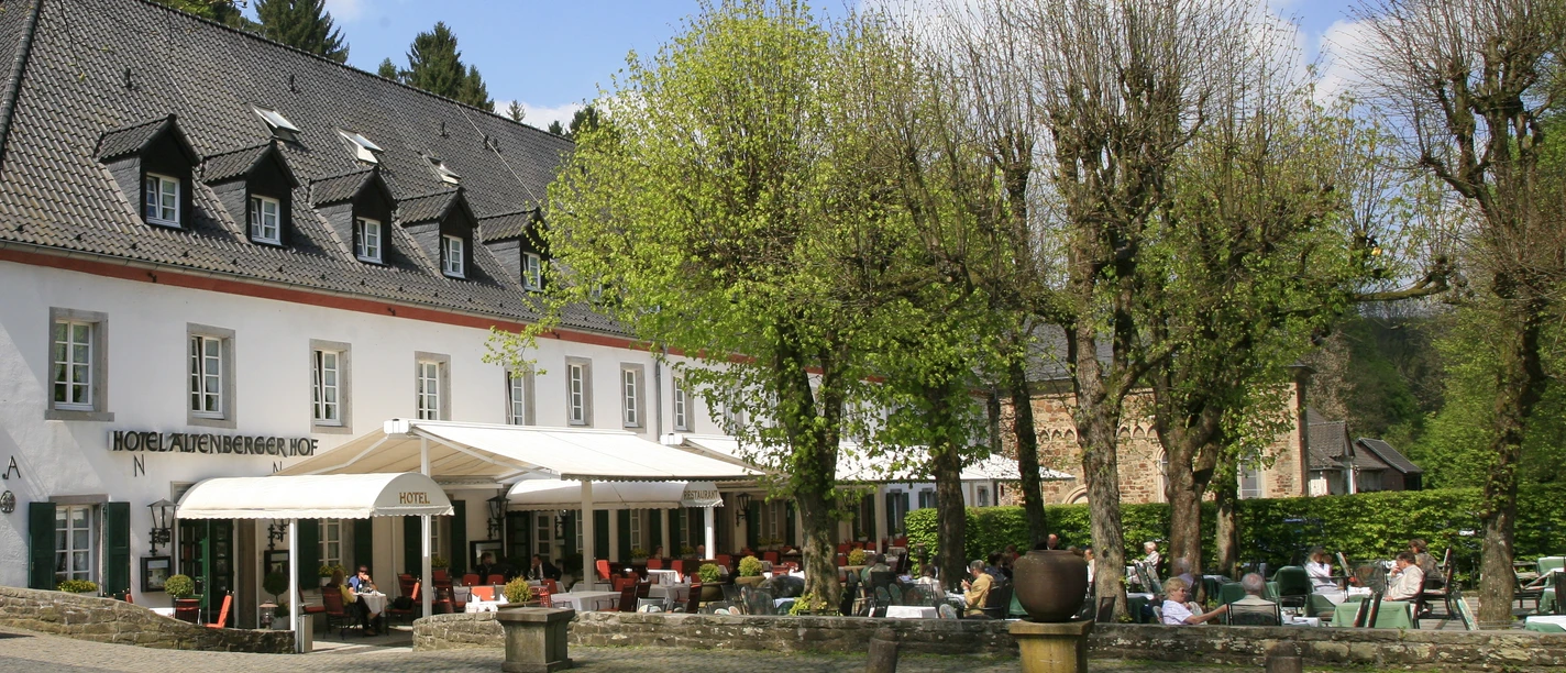 Altenberger Hof Ein charmantes Hotel mit Terrasse, umgeben von Bäumen, Gäste genießen die Sonne bei Getränken.