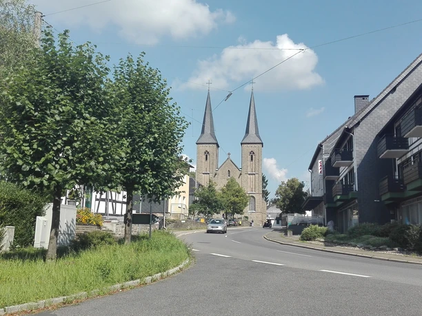 Overath Blick auf eine Straße mit einem Baum und einer Kirche im Hintergrund, flankiert von modernen Gebäuden.