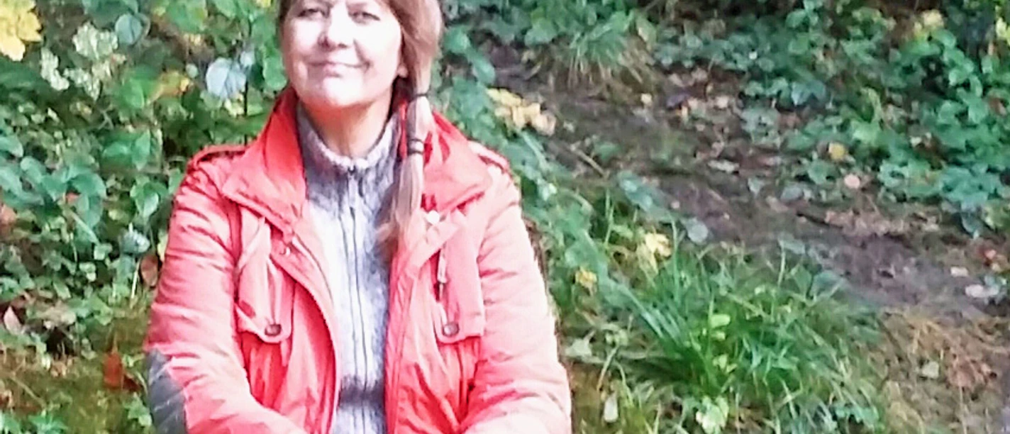 C. Dudda-Retzlaff.jpg Eine Frau in roter Jacke sitzt lächelnd auf einer Bank, umgeben von dichtem grünen Laub.