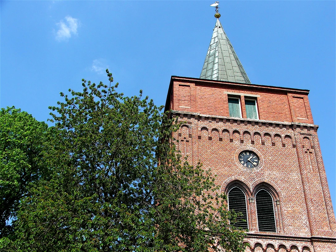Nikolaikirche Pewsum Roter Backsteinturm der Nikolaikirche Pewsum mit Uhr und Spitzdach vor blauem Himmel.
