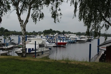 Boats are moored at jetties on a calm lake, framed by green trees and a cloudy sky.