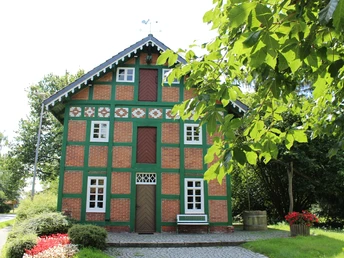 Rotes Fachwerkhaus mit grünem Gebälk, umgeben von Bäumen, mit Bank und Blumen vorne rechts.