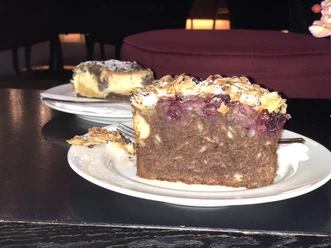 A slice of cake on a white plate, decorated with cherries and almonds on dark pastry.