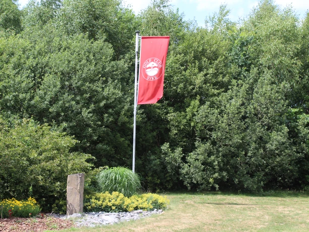 Een rode vlag van de Syke Golf Club wappert, omringd door groene struiken en gemanicuurde gazons.