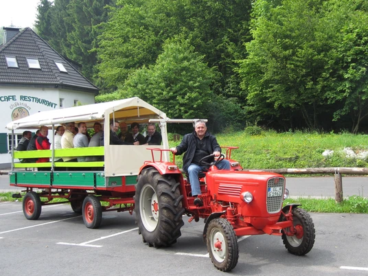Bauernhoftour mit dem Planwagen Gruppe von Menschen auf einem überdachten Anhänger, gezogen von einem roten Traktor, vor Waldkulisse.