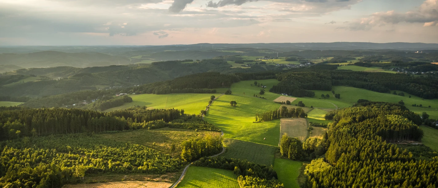 Reichshof Blockhaus Luftaufnahme einer grünen, hügeligen Landschaft bei Reichshof unter bewölktem Himmel im Abendlicht.