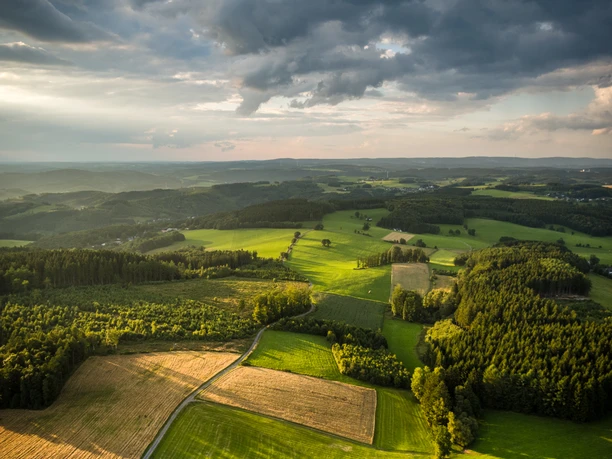 Reichshof Blockhaus Luftaufnahme einer grünen, hügeligen Landschaft bei Reichshof unter bewölktem Himmel im Abendlicht.