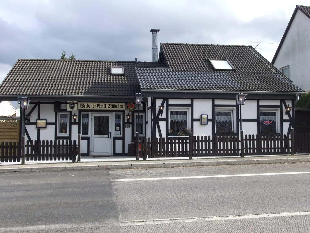 Weidener Grillstübchen.jpg Ein traditionelles Fachwerkhaus mit dunklen Balken und weißer Fassade, Holzzaun im Vordergrund.
