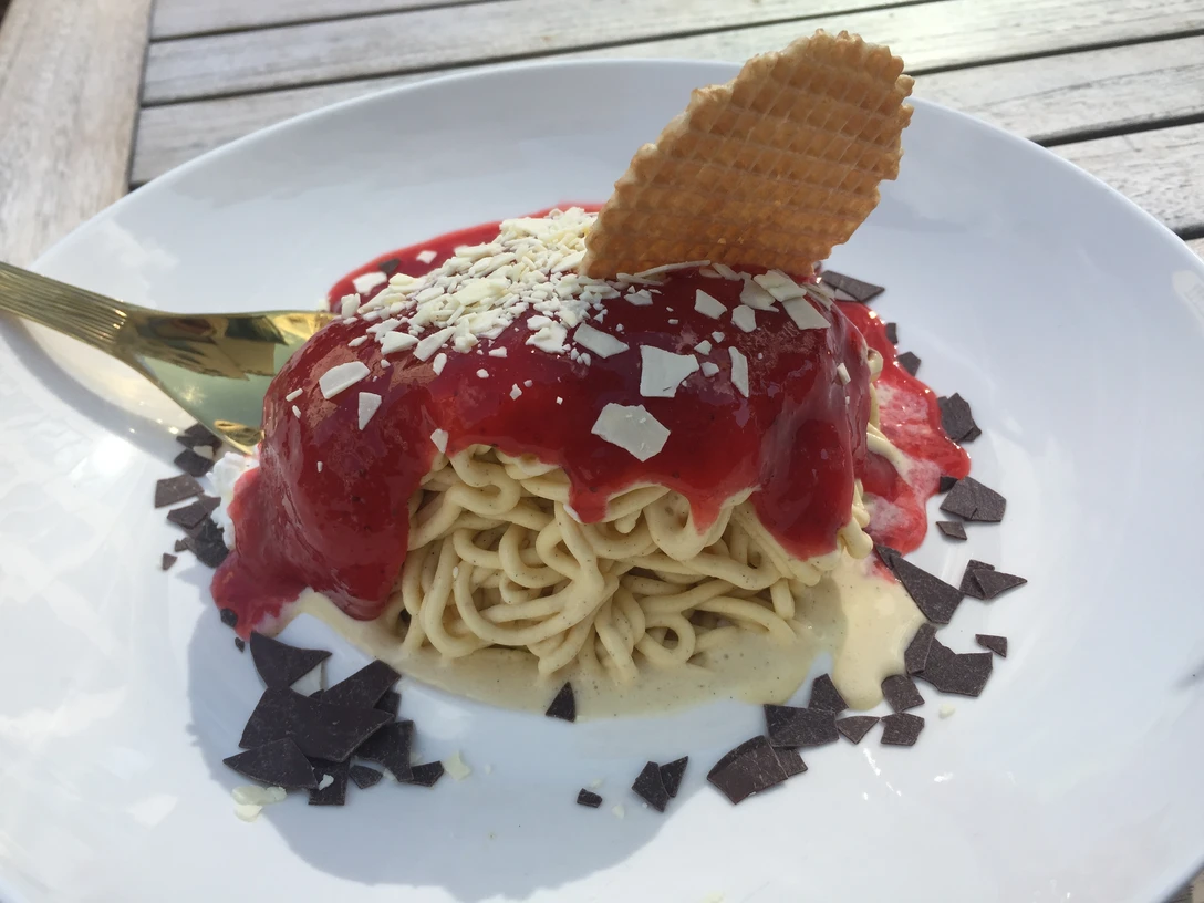 Spaghetti-Eis mit Erdbeersoße, weißer und dunkler Schokolade sowie einer Waffel auf einem Teller.