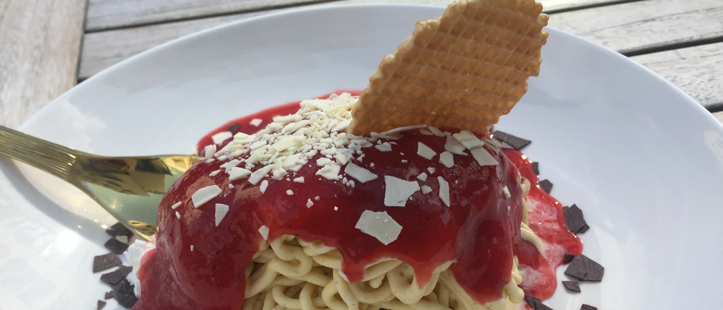 Spaghetti ice cream with strawberry sauce, white and dark chocolate and a waffle on a plate.