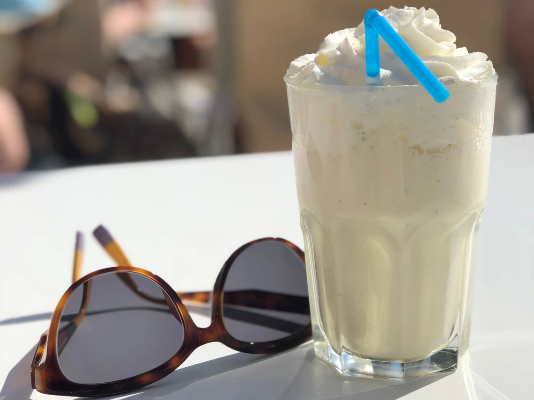 Ein erfrischender Milchshake mit Sahnehaube und blauem Strohhalm steht neben einer Sonnenbrille.