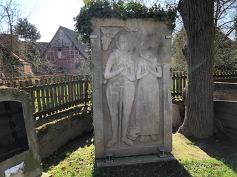 Kapelle Röhden Schlüsselburg Zwei antike Reliefs aus Stein, unter Efeuranken befestigt, stehen neben einem Baum mit Holzzaun.