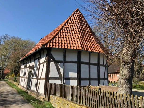 Historische Fachwerkkapelle mit rotem Ziegeldach, umgeben von Bäumen und Holzzaun, unter blauem Himmel.