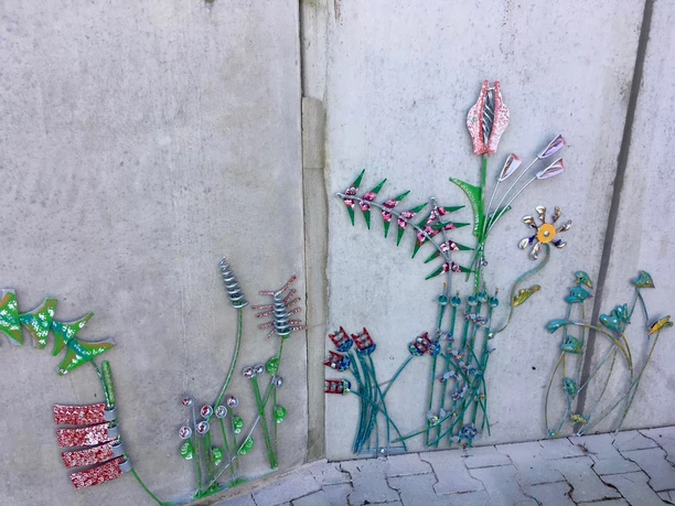 Lieblingsort Tange Ein gemustertes Betonwandbild zeigt farbenfrohe, abstrakte Pflanzen mit bunten Blüten und Blättern.