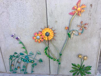 Blumenvielfalt am Lieblingsort Tange Eine Wand ziert ein farbenfrohes Kunstwerk mit fantasievoll gestalteten Metallblumen. Die Blüten strahlen in leuchtenden Orange- und Gelbtönen, ergänzt durch stilisierte grüne Blätter. Es verleiht der Wand einen lebhaften und kreativen Ausdruck.