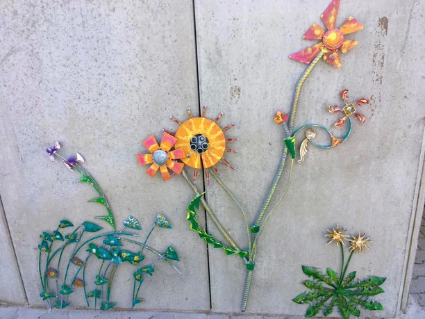 Blumenvielfalt am Lieblingsort Tange Eine Wand ziert ein farbenfrohes Kunstwerk mit fantasievoll gestalteten Metallblumen. Die Blüten strahlen in leuchtenden Orange- und Gelbtönen, ergänzt durch stilisierte grüne Blätter. Es verleiht der Wand einen lebhaften und kreativen Ausdruck.