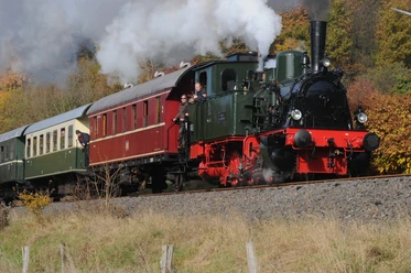 Symbolbild: Museumsbahn mit Dampflok Eine historische Dampflokomotive zieht bunte Waggons durch eine herbstliche Landschaft mit Rauchwolken.