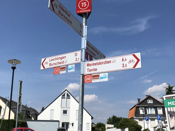 Raiffeisenplatz (Knotenpunkt 15) Wegweiser mit Rad- und Wanderwegekennzeichnung vor moderner Wohnsiedlung an der Straße bei sonnigem Wetter.
