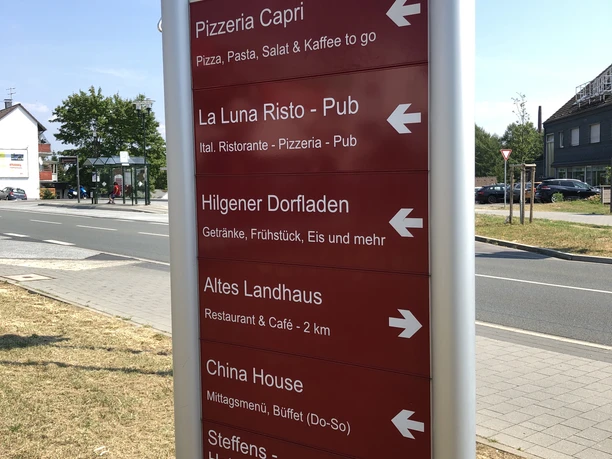 Gastro-Wegweiser (Haltestelle im Hintergrund) Wegweiser am Raiffeisenplatz in Burscheid-Hilgen mit Hinweisen zu lokalen Restaurants und Geschäften.