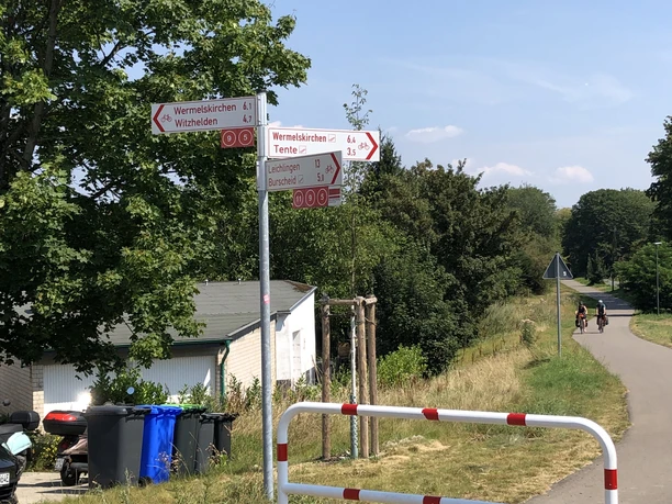 Einstieg Balkantrasse (Raiffeisenplatz) Zwei Verkehrsschilder mit Wanderweg-Hinweisen stehen an einem sonnigen Rad- und Wanderweg.