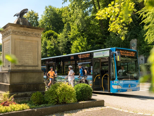 Bergischer FahrradBus, Haltestelle Hückeswagen Ein Bus fährt neben einem Denkmal mit Radfahrern im Grünen vorbei.