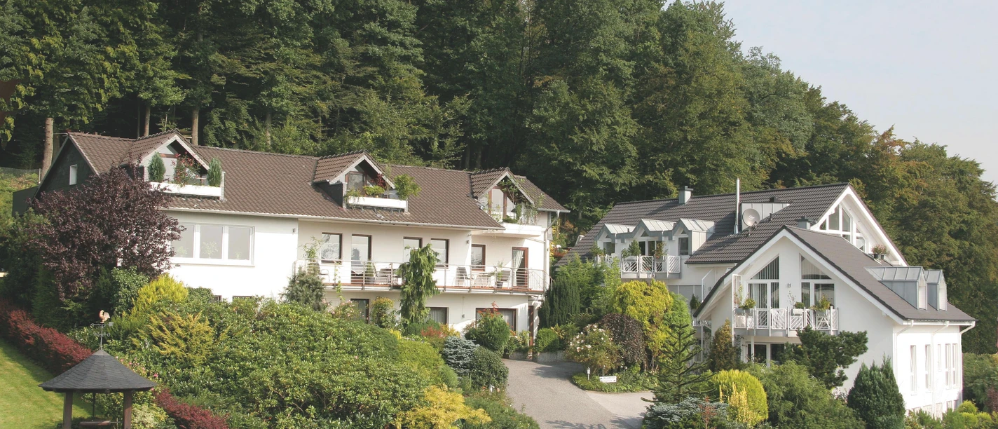 Zwei helle Pensiongebäude inmitten üppiger grüner Bäume und gepflegter Gärten an einem Hang.