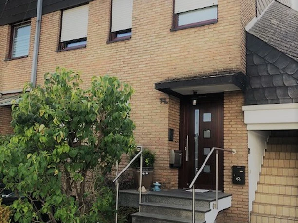 Frontansicht.jpg Vorderansicht eines zweistöckigen Hauses aus Ziegelstein, mit Treppe, Geländer und kleinen Garten.