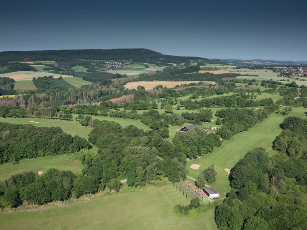 Weitläufige Golfplatzlandschaft inmitten sanfter Hügel, umgeben von dichten Wäldern und Feldern.