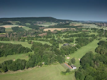 Golfplatz Weitläufige Golfplatzlandschaft inmitten sanfter Hügel, umgeben von dichten Wäldern und Feldern.