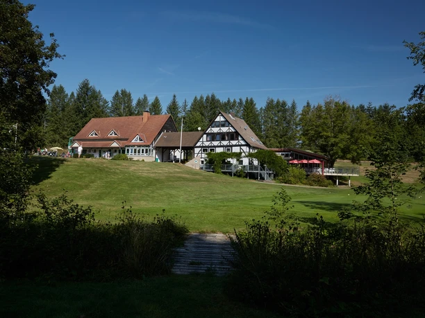 Das Clubhaus Clubhaus im Fachwerkstil mit weitläufigem Rasen, Bäumen im Hintergrund und klarem Himmel.
