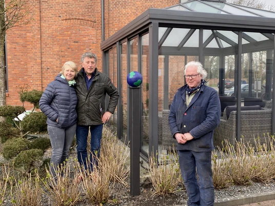 Lieblingsort Eisenhütte-Künstler Ridderbusch und Ehepaar Börjes Das Bild zeigt drei Personen vor einem roten Backsteingebäude. Links steht ein Mann in blauer Jacke neben einer Frau in grauer Jacke, die ihn umarmt. Rechts steht ein älterer Herr in blauer Arbeitsjacke, während ein kleiner schwarzer Hund zu seinen Füßen sitzt. Hinter ihnen ist ein modernes Gewächshaus an der Gebäudewand zu sehen.