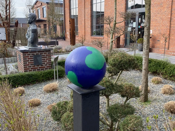Lieblingsort Eisenhütte Im Vordergrund des Bildes steht eine bunte Weltkugel aus zwei Farben auf einem Metallpfosten, umrahmt von gepflegten Gartenflächen und künstlerisch gestalteten Skulpturen vor einer historischen Backsteinfassade.
