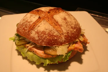 Ein belegtes Vollkornbrötchen mit frischem Salat, Gewürzgurke, Krautsalat und Schinken auf einem Teller.