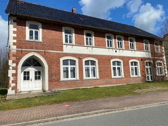 Zweistöckiges Backsteingebäude mit weißen Fenstern und Türen, umgeben von einer Wiese und Gehweg.