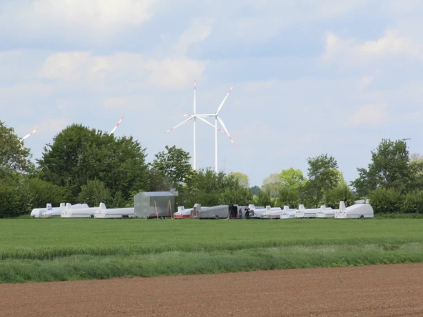 Gliders are parked on a green meadow; trees and a wind turbine can be seen in the background.