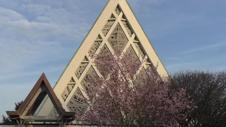 Kirche Eine Kirche mit modernem dreieckigem Dach und geometrischen Mustern, umgeben von blühenden Bäumen.