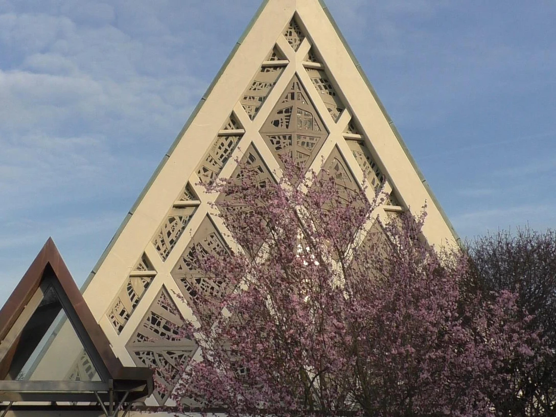 Kirche Eine Kirche mit modernem dreieckigem Dach und geometrischen Mustern, umgeben von blühenden Bäumen.