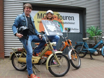 Zwei Personen lächeln auf Mofas vor einer Werbetafel für Mofa-Touren, ausgestattet mit Helmen.