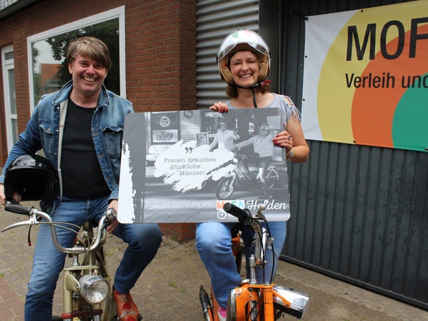 Two people on old mopeds are smiling; the woman is holding a humorous sign.