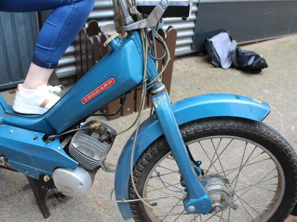 Ein blauer Zündapp-Mofa mit Reifenprofil, auf dem eine Person mit weißen Sneakern sitzt.