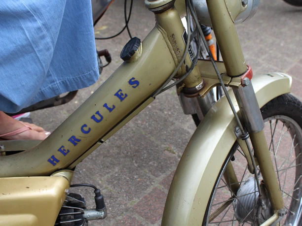 Ein goldenes Hercules Mofa mit blauem Schriftzug, seitlich, mit Vorderreifen und einem linken Bein.