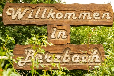 Ortsschild Berzbach.jpg Willkommensschild aus Holz, eingewachsen in grünen Pflanzen, mit der Aufschrift "Willkommen in Betzbach".