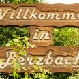 Ortsschild Berzbach.jpg Willkommensschild aus Holz, eingewachsen in grünen Pflanzen, mit der Aufschrift "Willkommen in Betzbach".