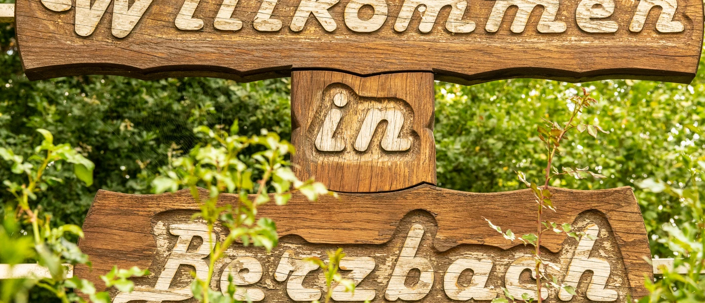 Ortsschild Berzbach.jpg Willkommensschild aus Holz, eingewachsen in grünen Pflanzen, mit der Aufschrift "Willkommen in Betzbach".