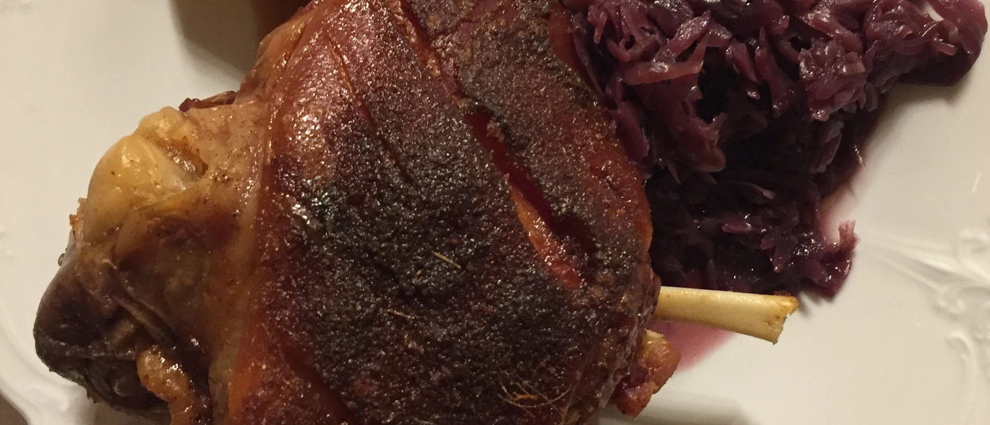 Crispy pork knuckle with a golden brown crust, served with potatoes and red cabbage on a white plate.