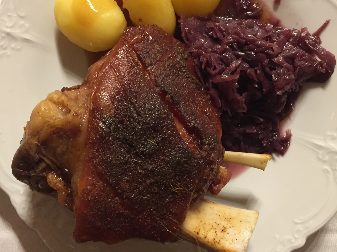 Haxe Knusprige Schweinshaxe mit goldbrauner Kruste, serviert mit Kartoffeln und Rotkohl auf weißem Teller.